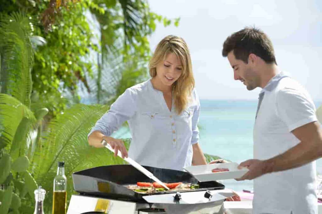 woman and man near grill grilling food on a beach with blue water in background, How to Grill on The Beach: Surprising [Simple Methods] To Prepare Food For Your Beach BBQ, can you grill on the beach, how to grill on the beach, can i grill on the beach, grill on the beach, can you cook on the beach, grilling on the beach, how to grill on the beach
