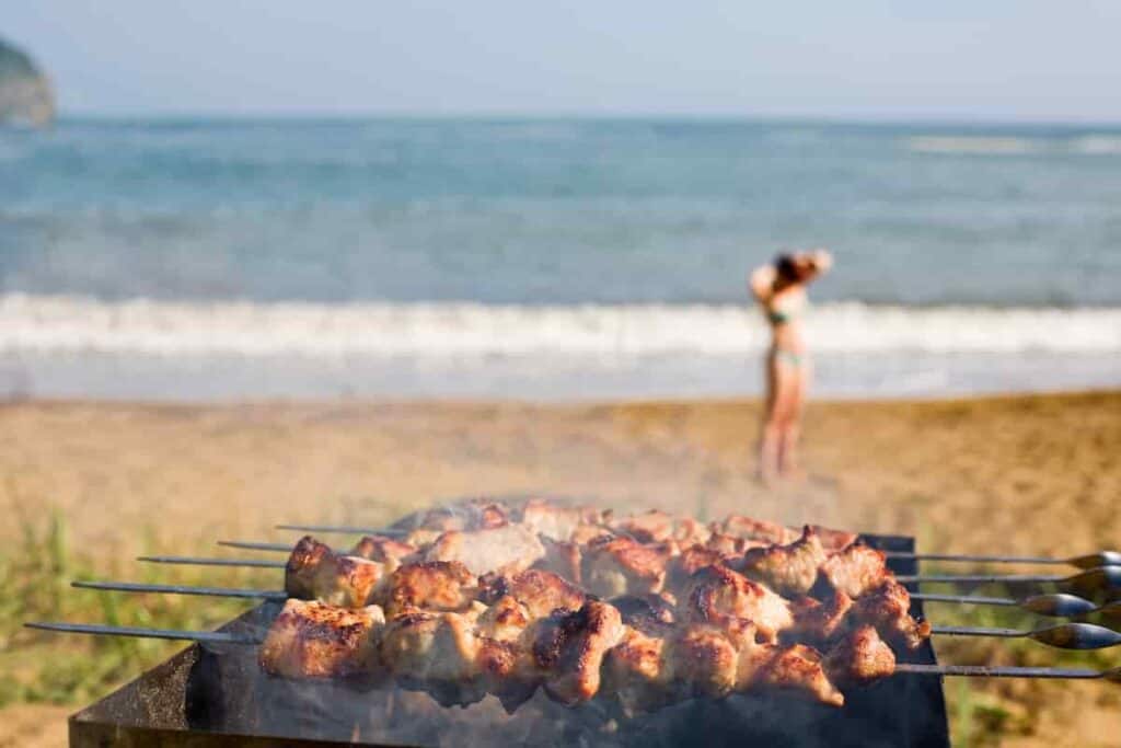 grill with shish kebabs grilling on a beach with girl and surf in the background, How to Grill on The Beach: Surprising [Simple Methods] To Prepare Food For Your Beach BBQ, how to grill on the beach, can you grill on the beach, grill on the beach, how to grill on the beach, grilling on the beach, can i grill on the beach, can you cook on the beach