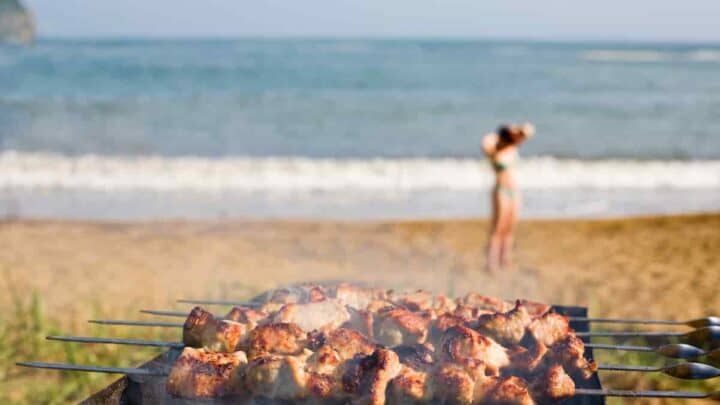 grill with shish kebabs grilling on a beach with girl and surf in the background, How to Grill on The Beach: Surprising [Simple Methods] To Prepare Food For Your Beach BBQ