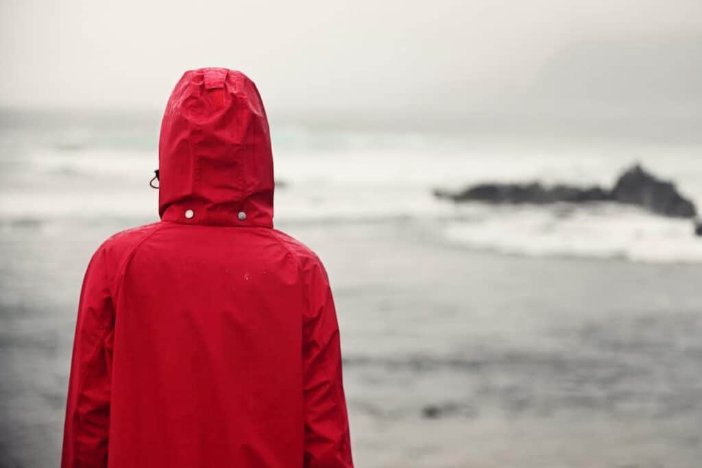 girl in red raincoat looking at the coast and surf on a rainy day, Making the Most of a Rainy Day at the Beach: Fun Rainy Day Activities to Enjoy Despite the Weather, what to do at the beach when it rains, rainy day beach activities, rainy day at the beach, rainy beach day, beach in the rain, rainy beach, rainy day beach activities
