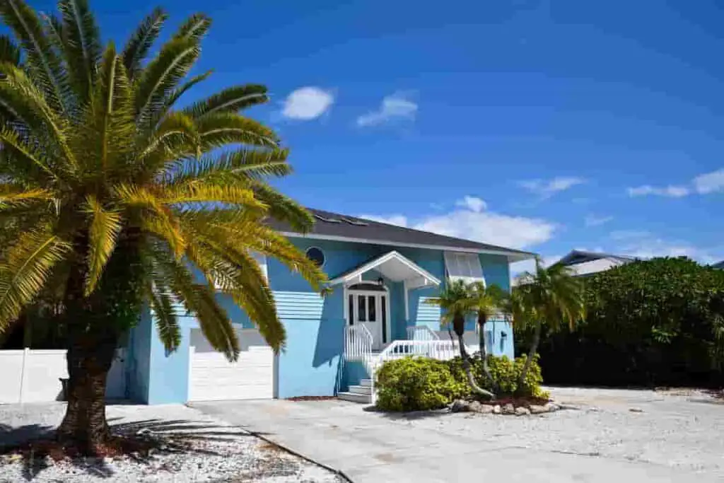 a blue beach house with a palm tree on the left in the front, How Much Does it Cost to Rent a Beach House? Renting vs. Buying: [Surprising Facts] Guide to Vacation Rentals, how much to rent a beach house, how much does a beach house cost to rent, how much does a beach house cost, how much is a beach house, how much are beach houses to rent, how much does it cost to rent a beach house for a week, how much is it to rent a beach house