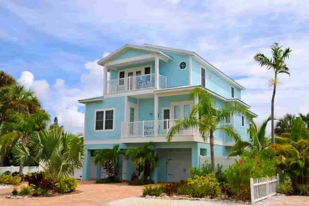 a three-story aqua blue beach house with blue sky and clouds in the background and palm tree on the right side, How Much Does it Cost to Rent a Beach House? Renting vs. Buying: [Surprising Facts] Guide to Vacation Rentals, how much does a beach house cost, how much is it to rent a beach house, how much to rent a beach house, how much does it cost to rent a beach house for a week, how much is a beach house, how much are beach houses to rent, how much does a beach house cost to rent