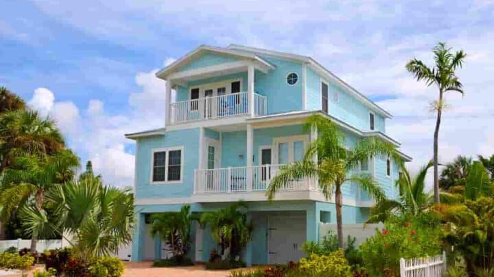a three-story aqua blue beach house with blue sky and clouds in the background and palm tree on the right side, How Much Does it Cost to Rent a Beach House? Renting vs. Buying: [Surprising Facts] Guide to Vacation Rentals