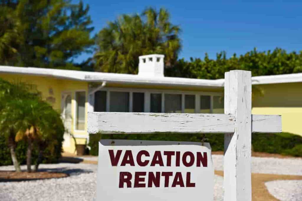 a sign that reads Vacation Rental with a yellow beach house in the background, How Much Does it Cost to Rent a Beach House? Renting vs. Buying: [Surprising Facts] Guide to Vacation Rentals, how much is it to rent a beach house, how much does it cost to rent a beach house for a week, how much does a beach house cost, how much does a beach house cost to rent, how much is a beach house, how much are beach houses to rent, how much to rent a beach house