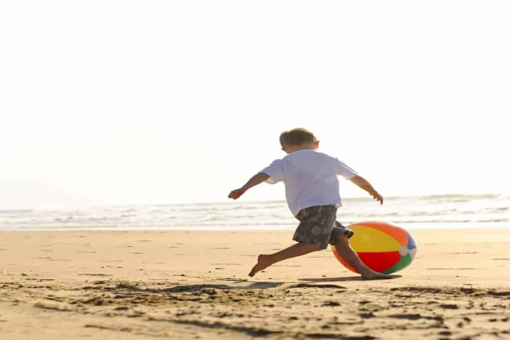 boy in white t-shirt running on the beach kicking a large colorful beachball, Top 9 Beach Ball Games for Fun at the Beach : Beach Ball Games For Kids