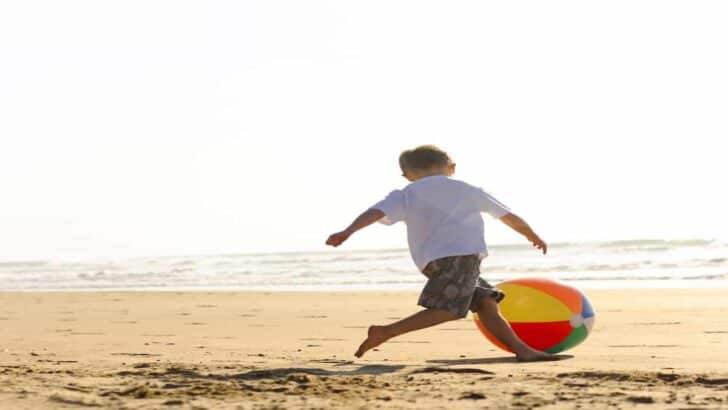 boy in white t-shirt running on the beach kicking a large colorful beachball, Top 9 Beach Ball Games for Fun at the Beach : Beach Ball Games For Kids