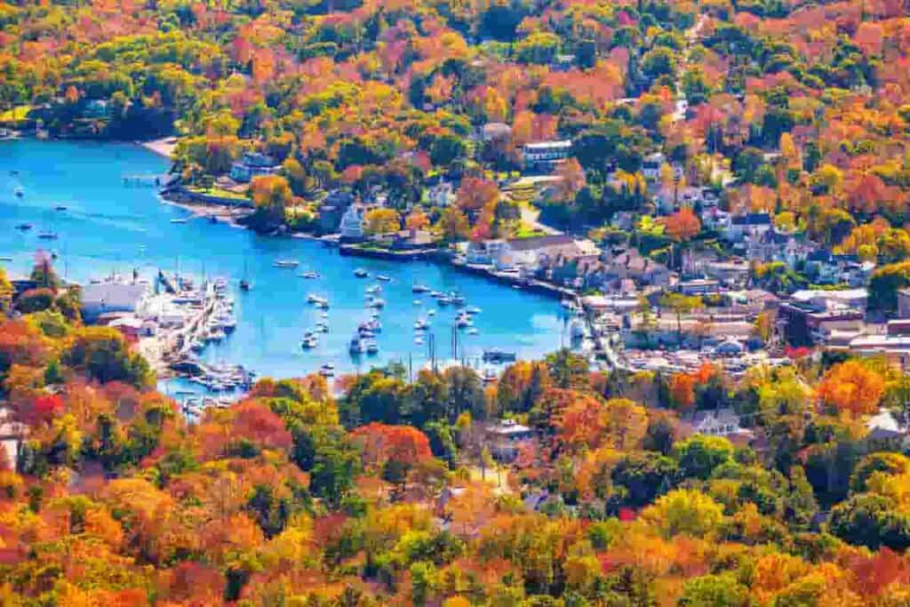 beautiful autumn colors surround a villiage and blue lake, 13 Prettiest Coastal Towns to Visit in Maine in Autumn-[Surprising Beautiful] Seaside, coastal towns to visit in maine in autumn, best places to visit in maine in the fall, coastal towns in maine, best coastal towns in maine, maine coastal towns, best beach towns in maine, places to visit in maine in the fall, best towns in maine to visit in fall