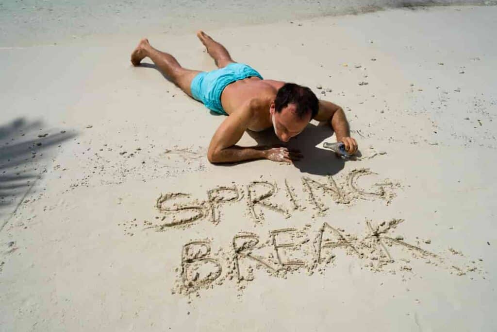 man lying down on his belly overlooking the message spring break written in the sand, Top 15 Best Beach Vacation for College Students [Spring Break Destinations], beach vacation for college students, beach vacation spots, spring break destinations for college students, best spring break destinations for college students, best beaches for college students, cheap college student trips, spring break destinations, vacations for college students