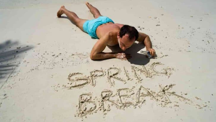 man lying down on his belly overlooking the message spring break written in the sand, Top 15 Best Beach Vacation for College Students [Spring Break Destinations]