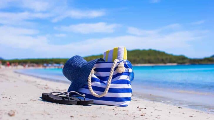 A blue and white striped tote bag on the sand at the beach, 11 Beach Travel Essentials to Pack – Minimalist Approach [Plus Packing List], Minimalist Beach Packing, UV-Protection Essentials, Beach Footwear Choices, Water-Resistant Tech Gear, essential beach items, minimalist packing for beach vacation, beach trip essentials