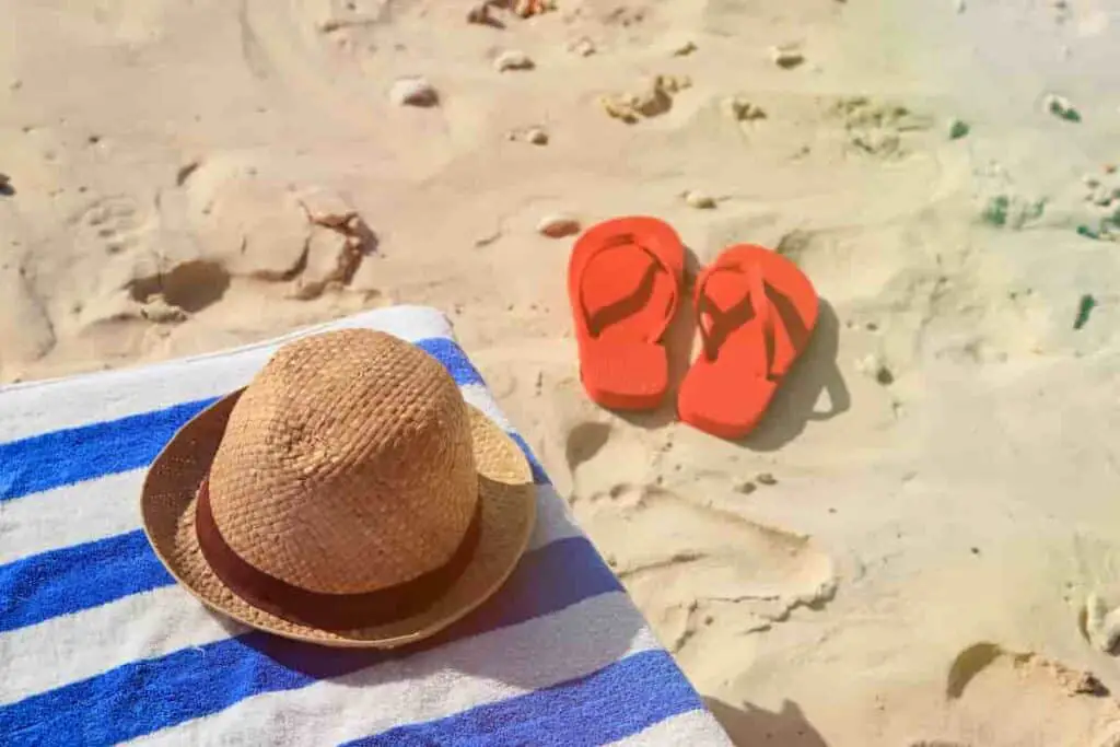 A blue and white striped towel with a hat on the sand with red flip flops next to it, 11 Beach Travel Essentials to Pack – Minimalist Approach [Plus Packing List], Water-Resistant Tech Gear, minimalist packing for beach vacation, Minimalist Beach Packing, Beach Footwear Choices, UV-Protection Essentials, essential beach items, beach trip essentials