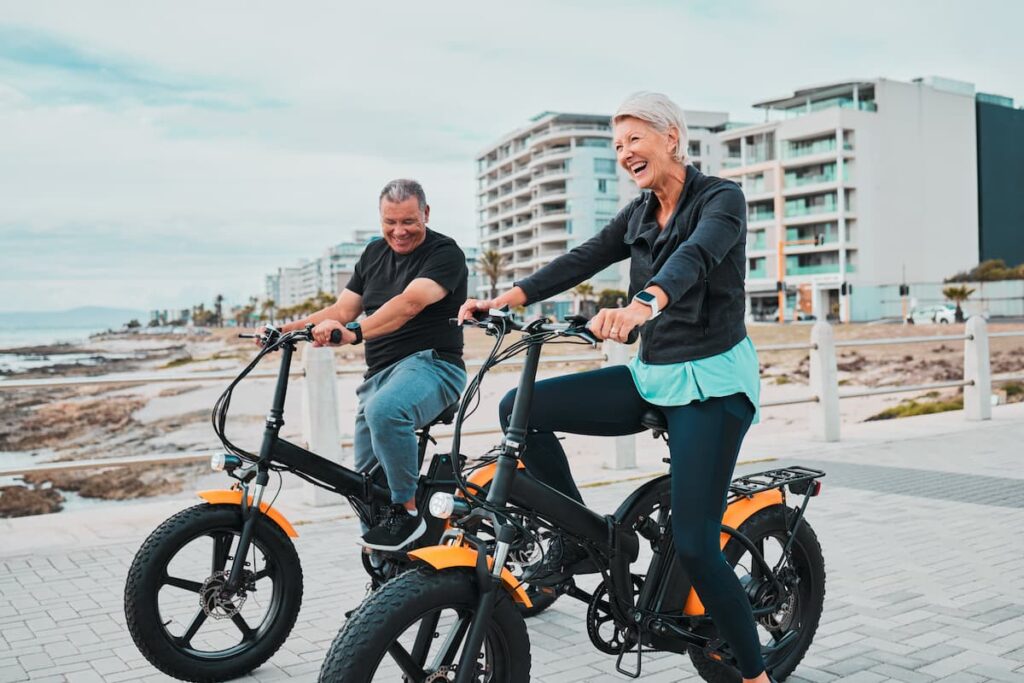 elderly man and woman on beach bikes smiling with buildings in the background, Best Beach Towns in the World to Retire - Surprising Towns, Retirement Criteria, Affordability, Activities, best beach towns to retire, most affordable beach towns to retire in the world, best beaches to retire in the world, cheapest beach towns to retire in the world