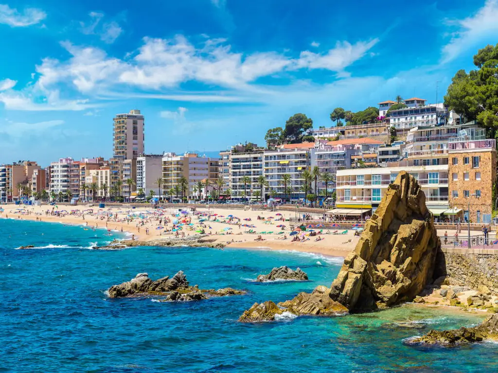 beach photo of Spanish beach with people on beach and blue water with beach town buildings in background, Towns Near Barcelona With A Beach: 11 Surprising Towns [With Beaches], cities near barcelona, barcelona beach resorts, best beach towns near barcelona, barcelona beach, beach resorts near barcelona, best coastal towns near barcelona, beach towns near Barcelona