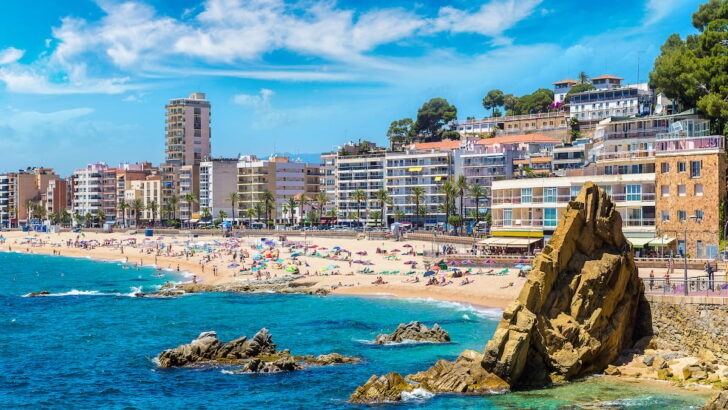 beach photo of Spanish beach with people on beach and blue water with beach town buildings in background, 2024 Best Towns Near Barcelona With A Beach: 15 Tops Spots