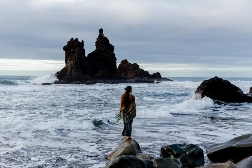 Unrecognizable female tourist standing on rocky coast, 9 [Best] Things to Do at the Beach When it’s Cloudy: A Gloomy Day Turned Into Fun, overcast beach, what to do on a cloudy day, cloudy beach day, cloudy day at the beach, activities during cloudy day, cloudy beach, beach on a cloudy day