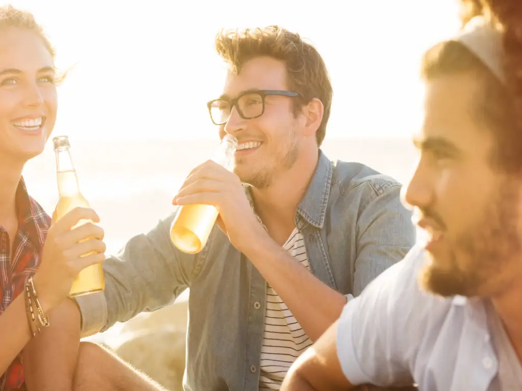 man with friends sitting on beach with bottle in hand, 21 [Best] Beach Photo Ideas for Guys- Poses Make The Difference, beach poses for men, Mens Beach Poses, beach photo ideas for guys, beach pictures poses man, Action Shots on Beach, beach pose ideas, beach poses man