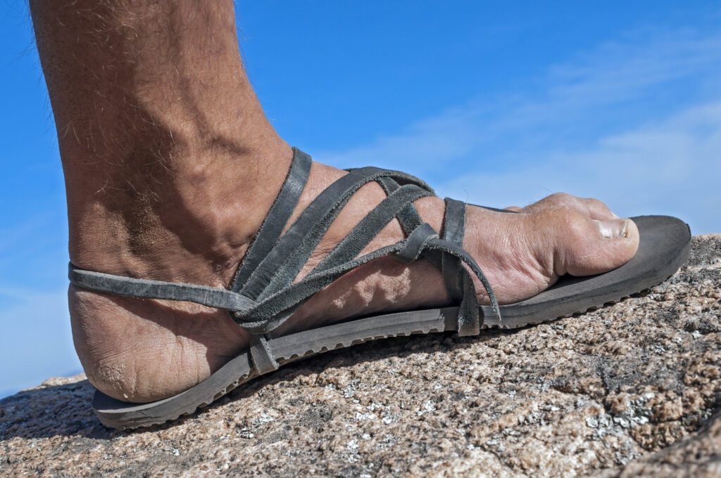 One black leather sandal worn by man standing on rock, active beach hiking footwear Experience Comfort: Unveiling the Best Beach Sandals For Men, best beach footwear, beach sandals for men, best sandals for men, men's sandals, beach thong sandals for men, active beach footwear, beach sandals for sand, water beach sandals, hiking beach sandals,