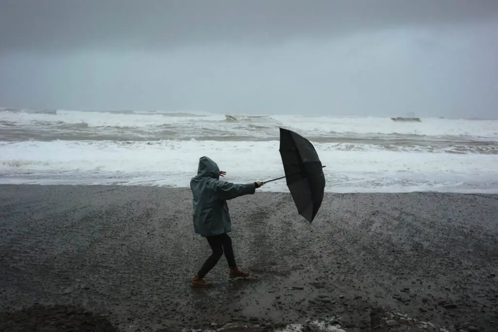 Full body of anonymous person in hood standing in wind with umbrella on sandy shore near stormy sea in rainy weather, Making the Most of a Rainy Day at the Beach: Fun Rainy Day Activities to Enjoy Despite the Weather, what to do at the beach when it rains, rainy day beach activities, rainy day at the beach, rainy beach day, beach in the rain, rainy beach, rainy day beach activities